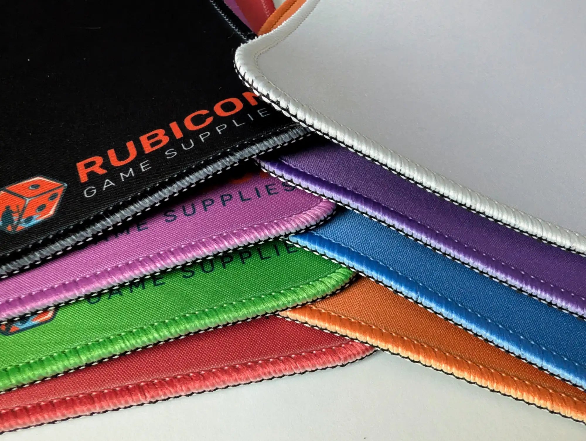 A stack of colorful, stitched-edge gaming mat pads in vibrant hues including black, purple, blue, green, and orange.